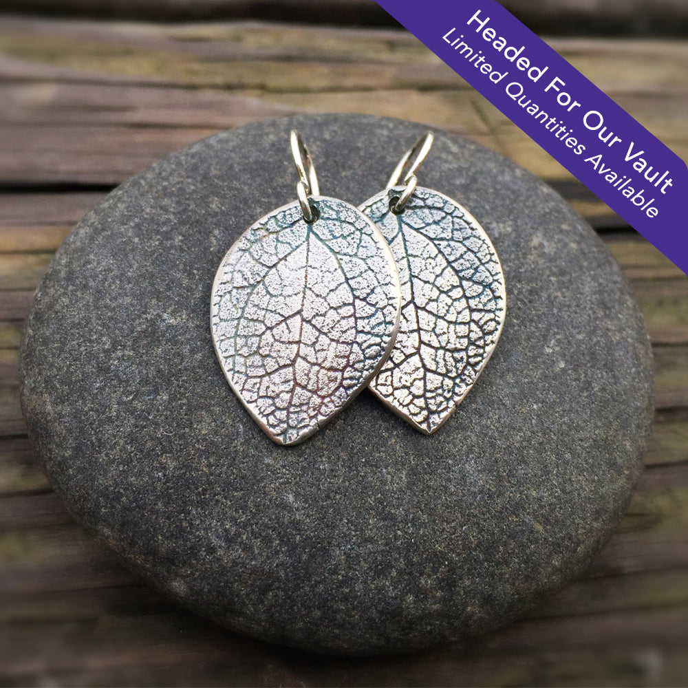 "Headed For Our Vault. Limited Quantities Available" oxidized silver salal leaf earrings on top of round stone on wood grain background
