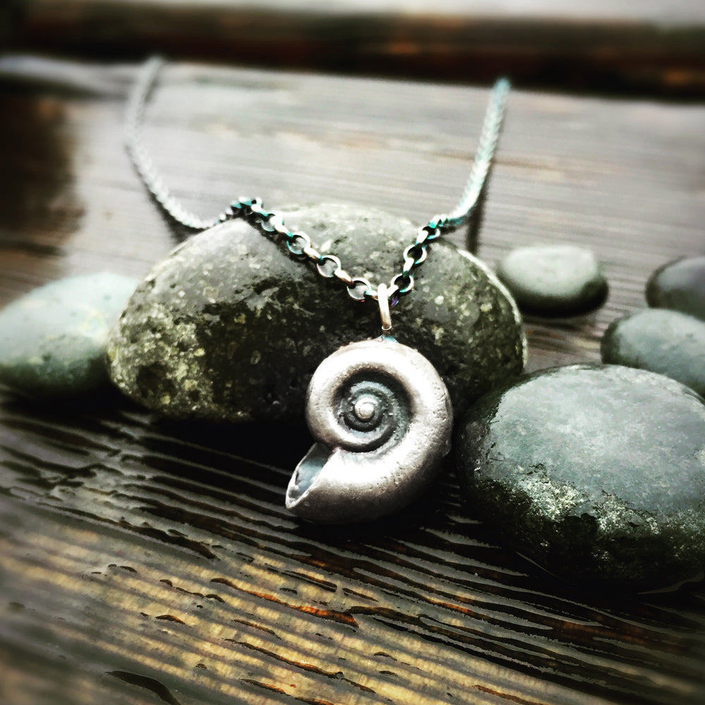 oxidized silver nautilus pendant necklace perched against some rocks on a rough wood grain backdrop 