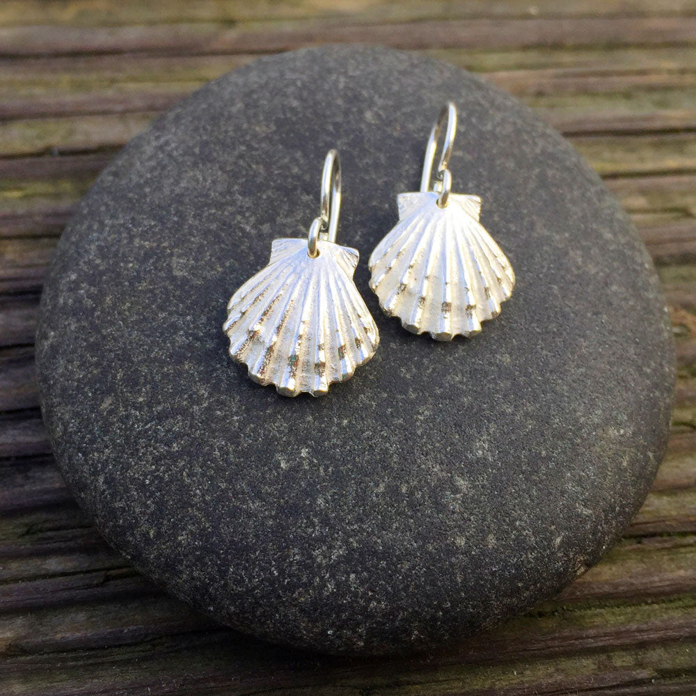 sterling silver scallop shell earrings perched on top of a round rock on a wood grain background