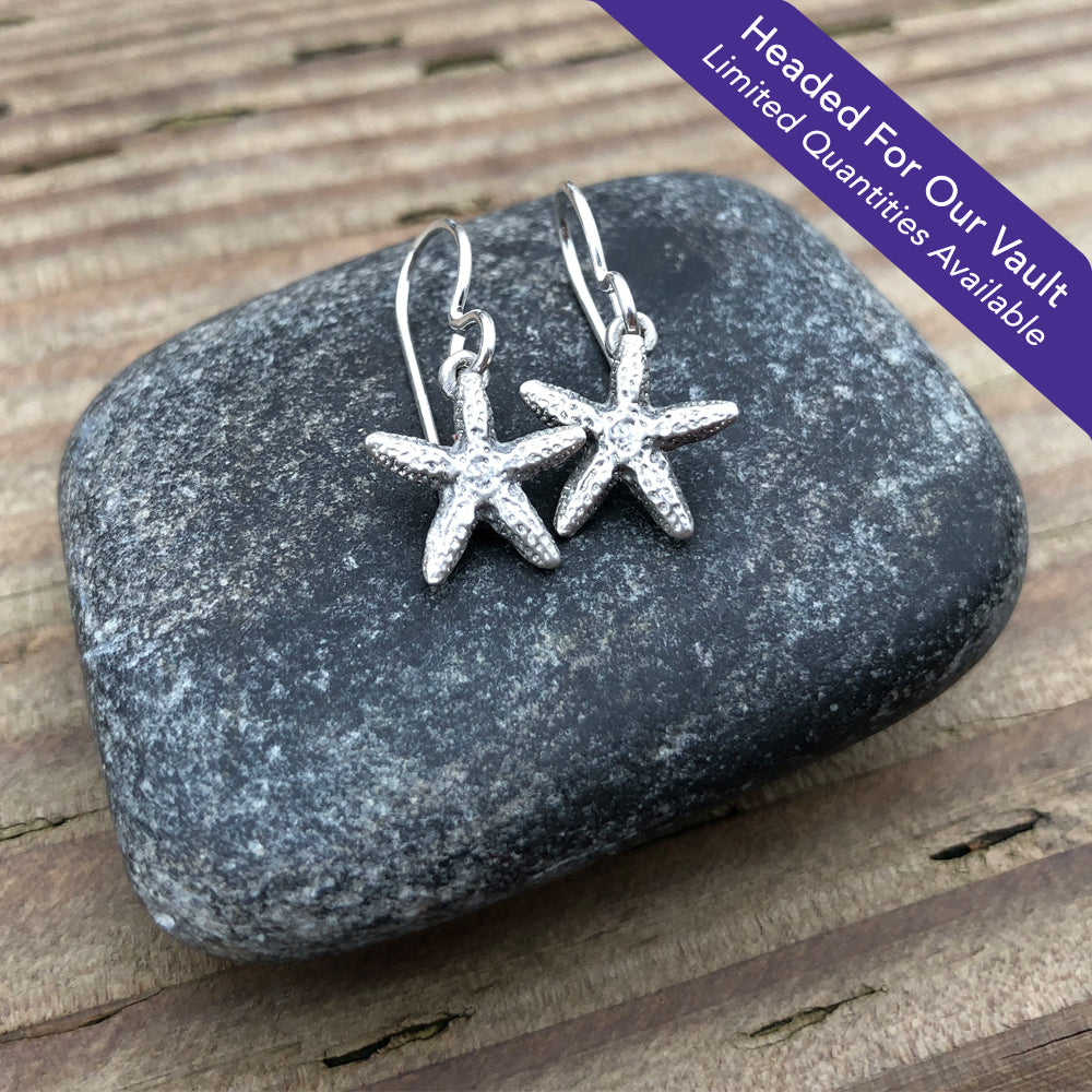 "Headed For Our Vault. Limited Quantities Available" small oxidized silver starfish earrings on a stone with a wood grain background
