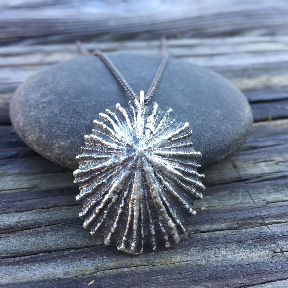 beautifully detailed oxidized silver limpet necklace, leaning against a round stone on a wooden board